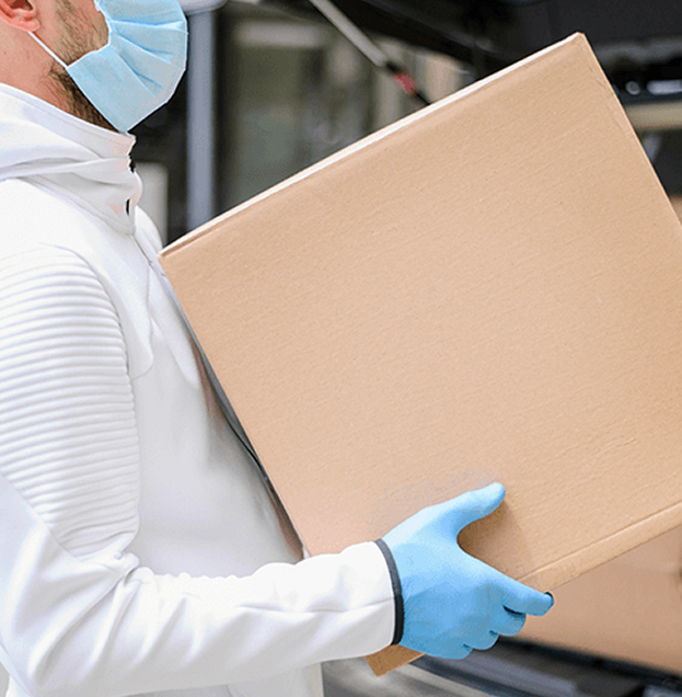 Warehouse worker in a mask and gloves carrying a sealed shipping carton